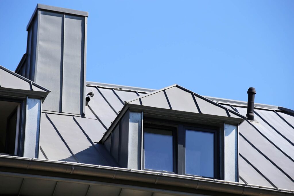 A rooftop with metallic cladding features dormer windows and a chimney. The metal has a reflective, silvery finish, and the sky above is clear and blue.