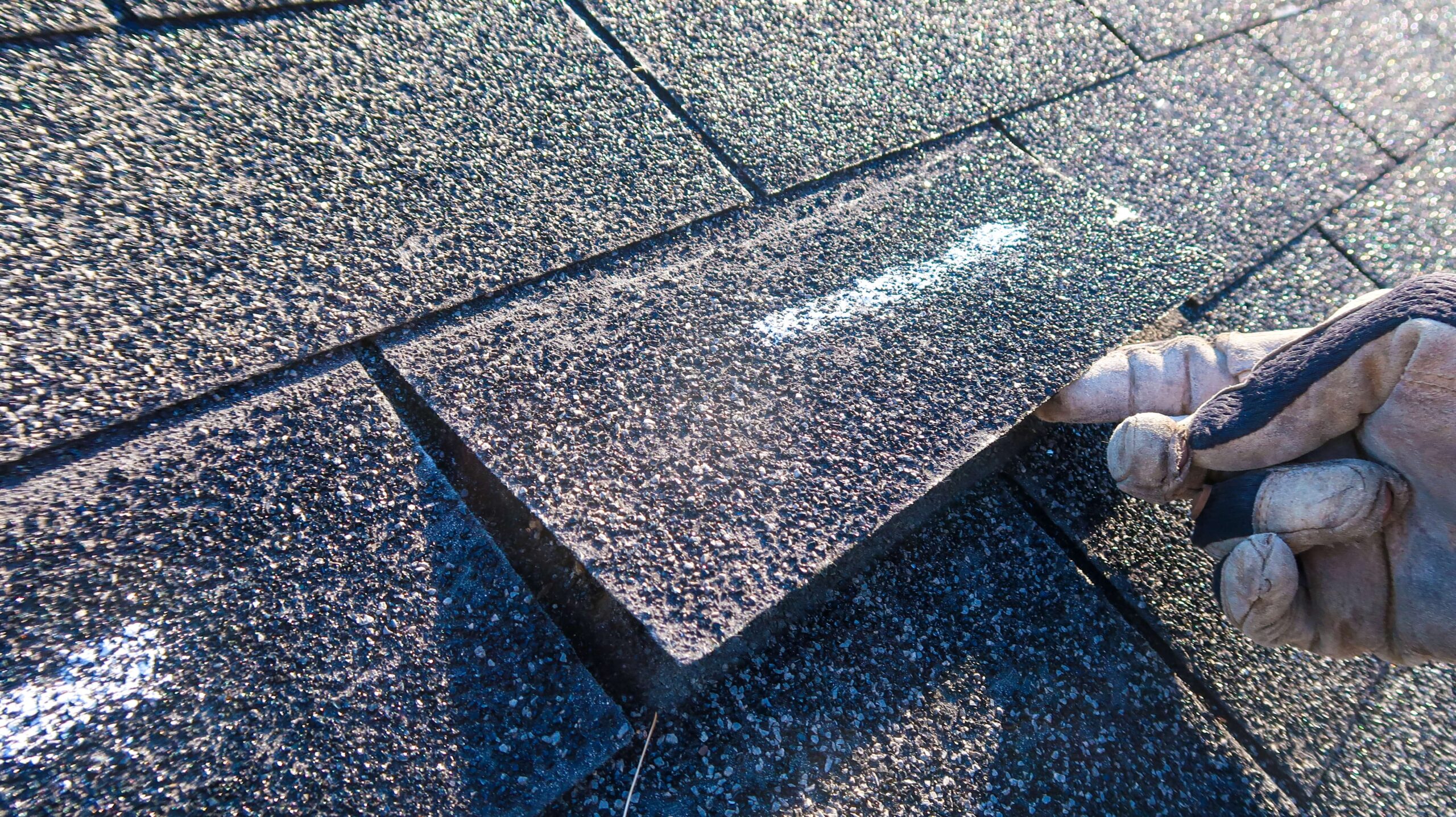 A person wearing a glove is lifting a roof shingle to check for damage on a shingled rooftop. The shingles are dark-colored and textured, and the scene is lit by natural sunlight.