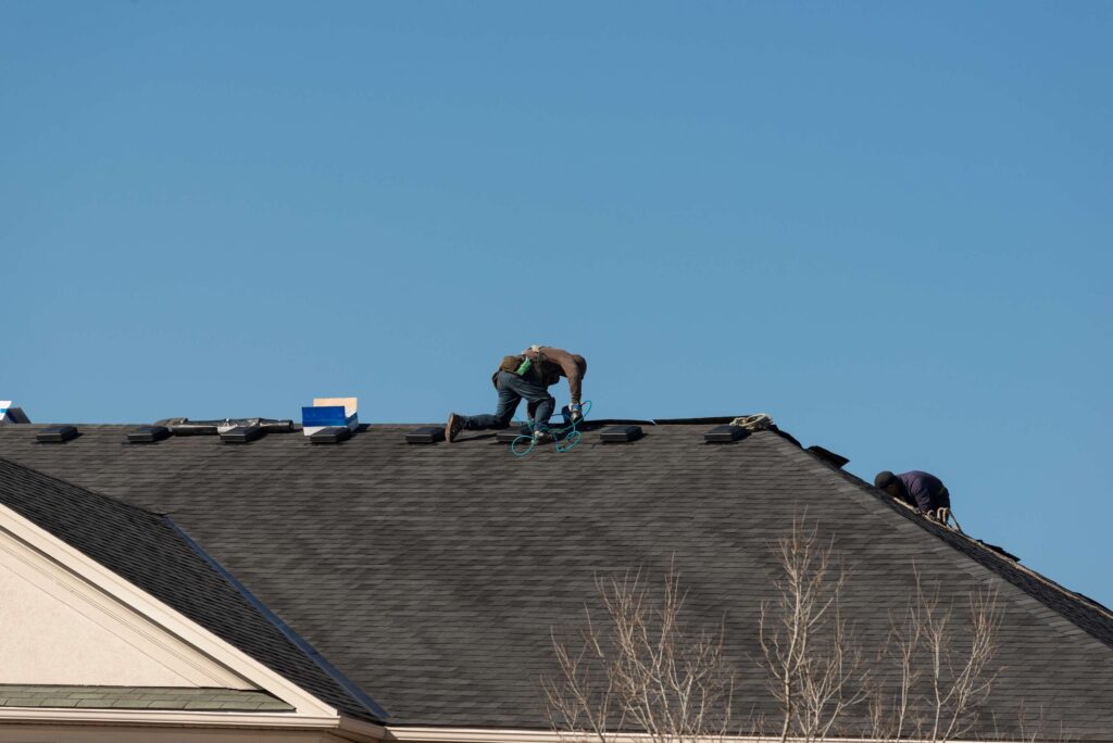 Two workers from a New Orleans Roofing Company are on a house roof under a clear blue sky, installing or repairing shingles. One kneels in the center with tools, while the other is near the edge. Equipment and a blue cooler sit nearby.