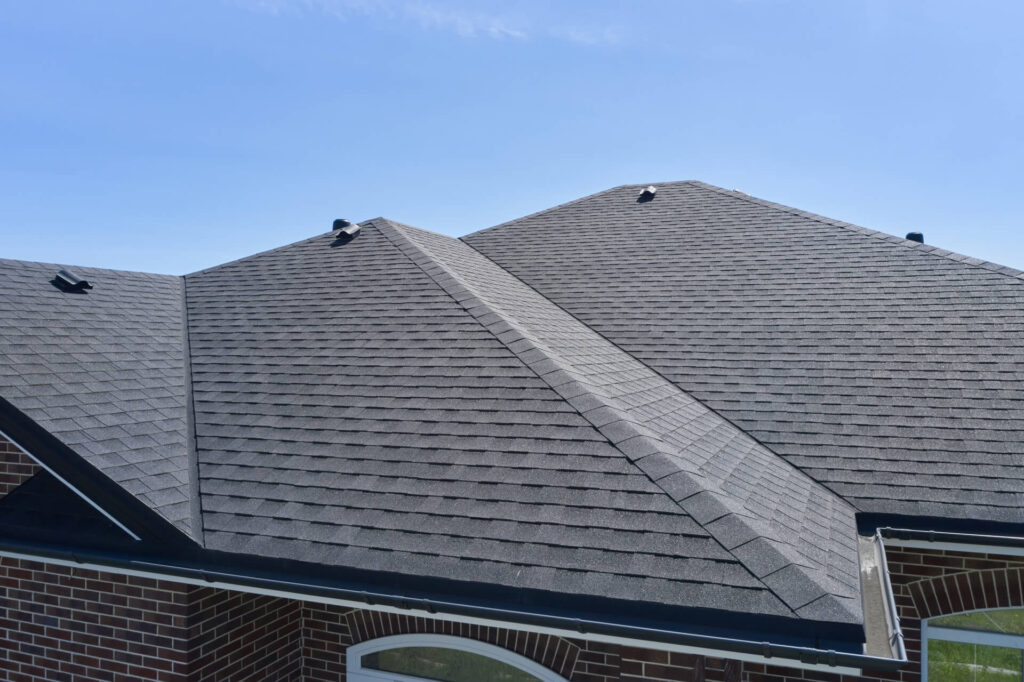 Gray shingled roof with multiple peaks against a clear blue sky. The brick building corner is slightly visible at the bottom.