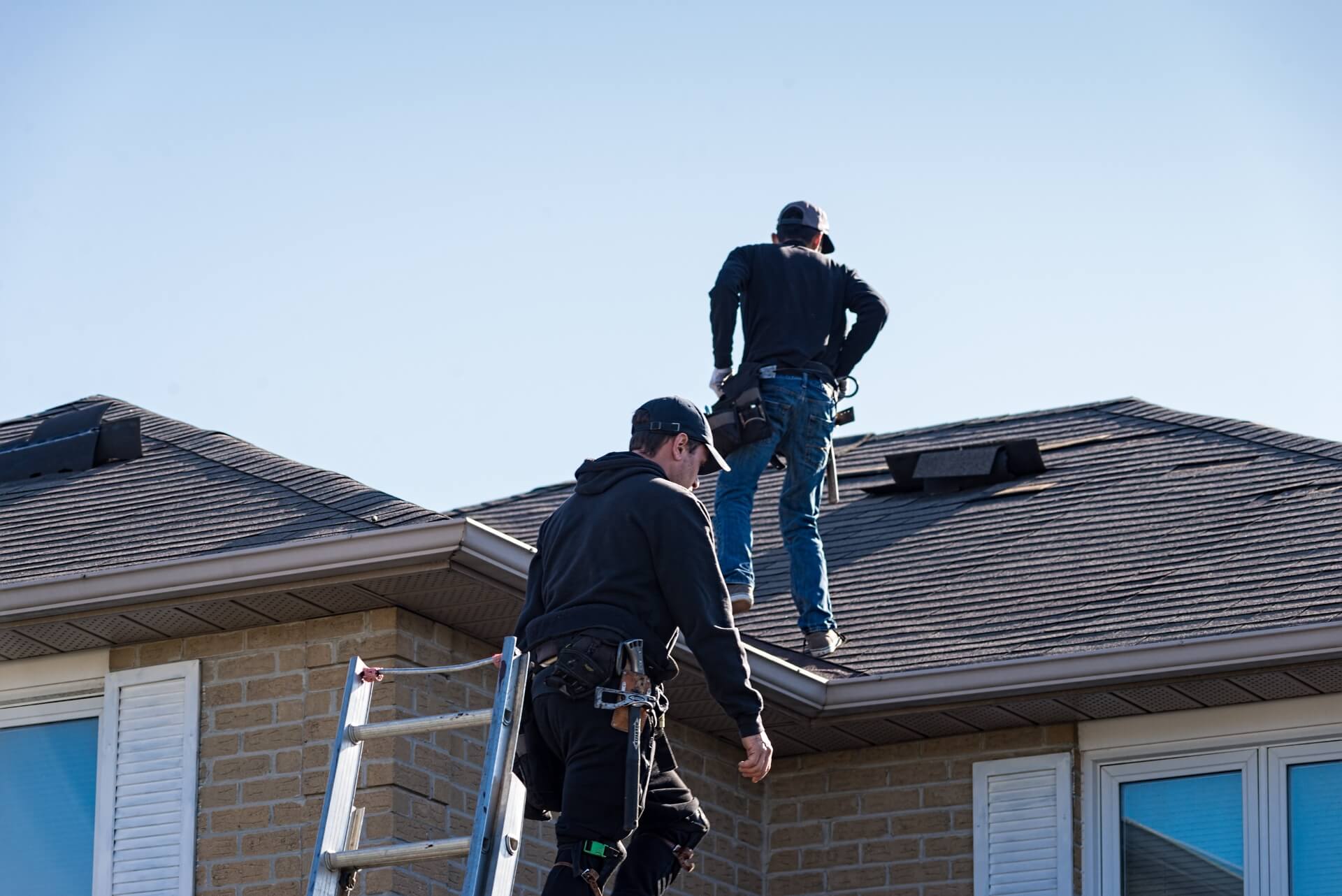 Two workers are standing on a roof, one near a ladder and the other further up. They are wearing casual clothes and caps, handling roof maintenance or repair tasks. The sky is clear, and the building is made of light-colored bricks.