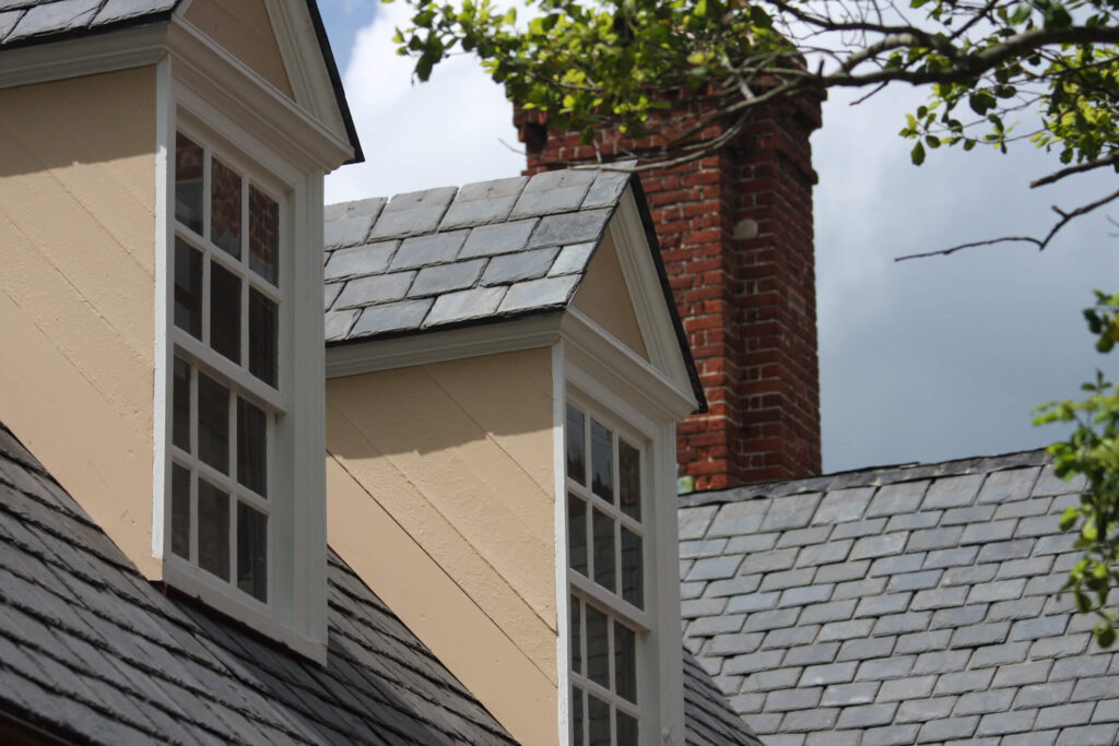 Gabled windows with white trim are seen on a slate-roofed house. A red brick chimney stands between the windows. Tree branches with green leaves frame the scene above against a cloudy sky.