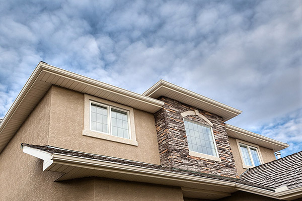 Close-up view of a modern house exterior with beige stucco walls, stone accents, white trim, large windows, and Cedar Shake Shingles on the roof, set against a partly cloudy sky.