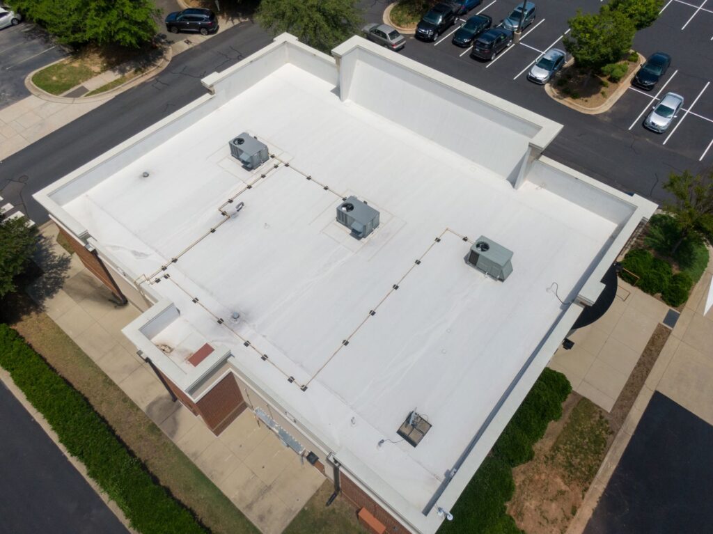 Aerial view of a flat white commercial building roof with HVAC units and pipes, showcasing Commercial Roof Coating New Orleans, surrounded by sidewalks, landscaping, and a parking lot with several parked cars.