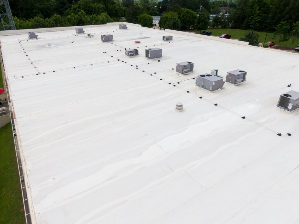 Aerial view of a large, flat white commercial roofing New Orleans building with several HVAC units and vents evenly spaced across the surface, surrounded by green trees and a parking lot.