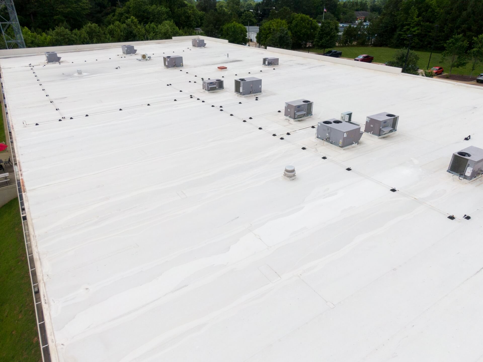 Aerial view of a large, flat white commercial roofing New Orleans building with several HVAC units and vents evenly spaced across the surface, surrounded by green trees and a parking lot.