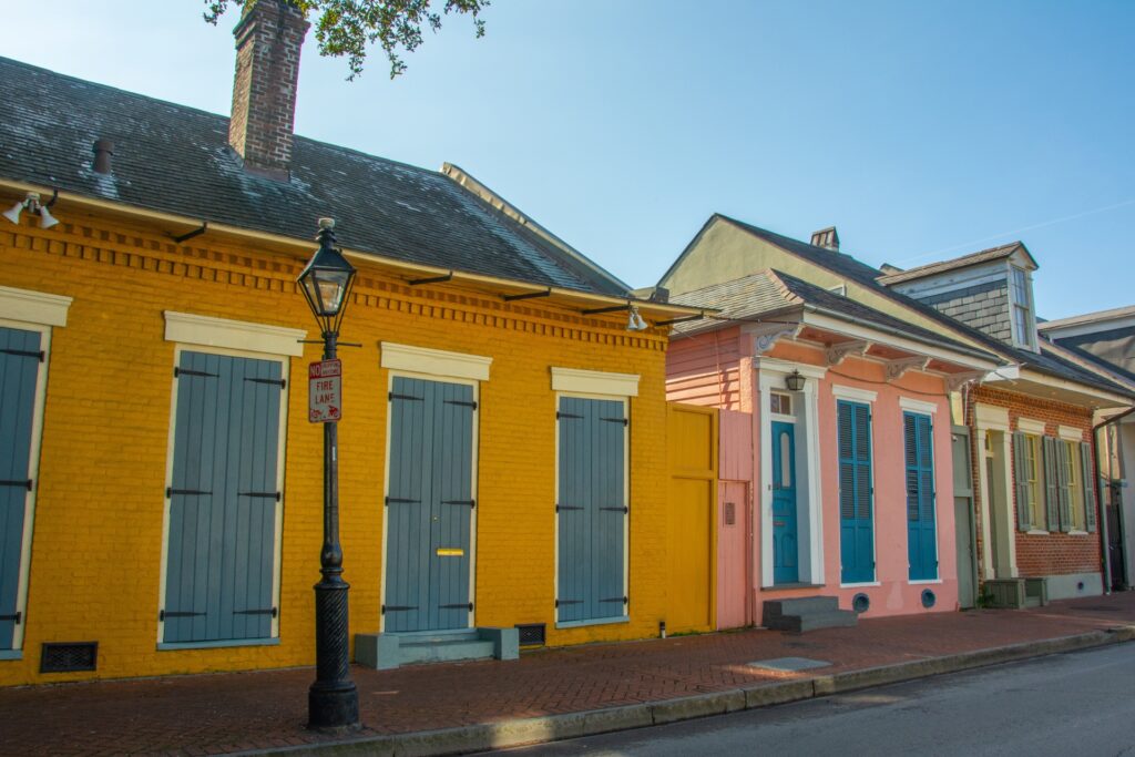 A vibrant street scene shows yellow and pink houses with blue shutters and doors, fresh from a roof replacement New Orleans locals trust, with a brick sidewalk and a vintage streetlamp under a clear blue sky.
