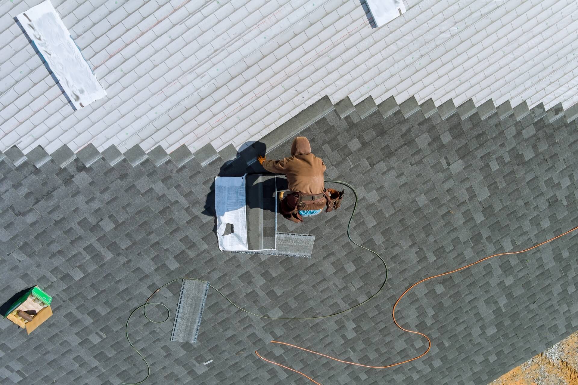 A person in a brown hoodie works on a sloped roof, installing vent covers or shingles—a typical scene in Residential Roofing New Orleans. Tools, metal pieces, and an orange extension cord are scattered nearby. The roof has two shades of shingles.