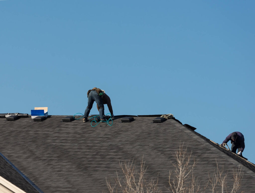 Two workers are installing shingles on the roof of a house under a clear blue sky. Specializing in residential roofing New Orleans, they use their tools and equipment, with one near the ridge and the other closer to the edge.