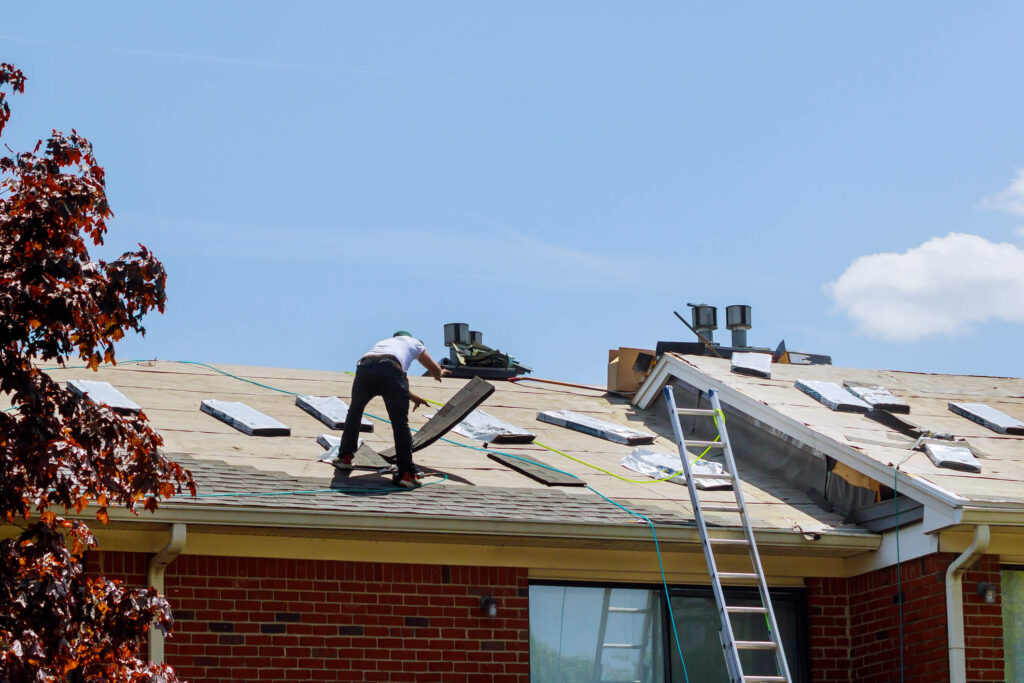 A worker installs shingles on the roof of a brick building under clear blue skies. Tools and roofing materials for residential roofing in New Orleans are scattered across the roof, and a ladder leads up from the ground.