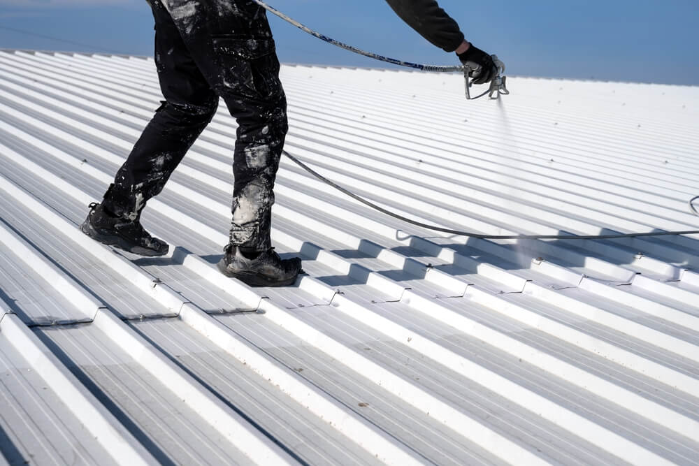 Person in black workwear spray painting a white corrugated metal roof. The worker is standing on the roof, holding a spray gun connected to a hose, with visible paint splatters on clothing. The sky is clear in the background.
