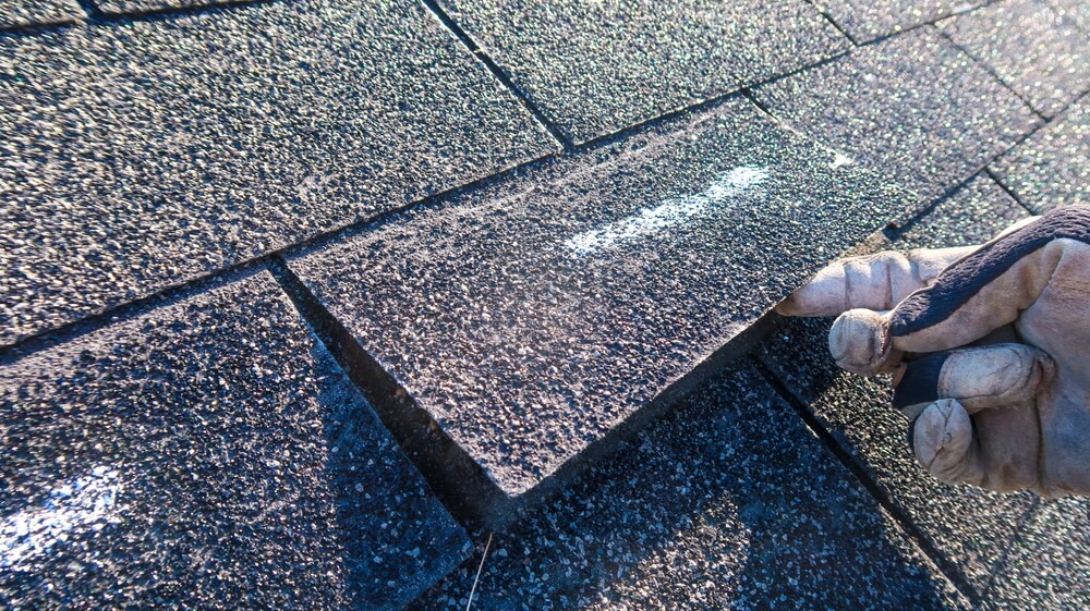 Close-up of a hand in a work glove lifting a shingle on a rooftop. The asphalt shingles are textured and dark-colored, with sunlight casting shadows. The focus is on the single lifted shingle and the gloved hand.